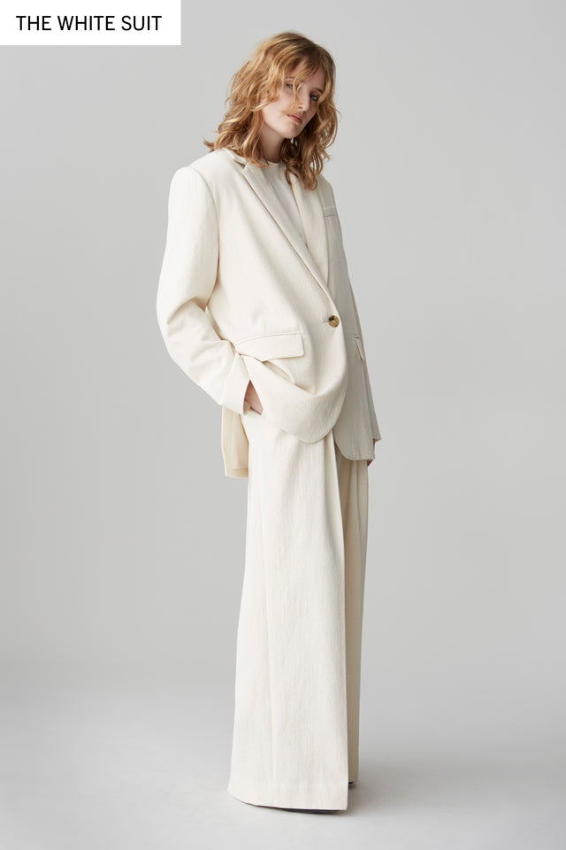 A woman wearing The Summer Suit by BLMBRG — a white cotton-wool tailored jacket with wide-leg trousers, styled with a white T-shirt. A timeless women’s suit, part of the summer edit (£595).