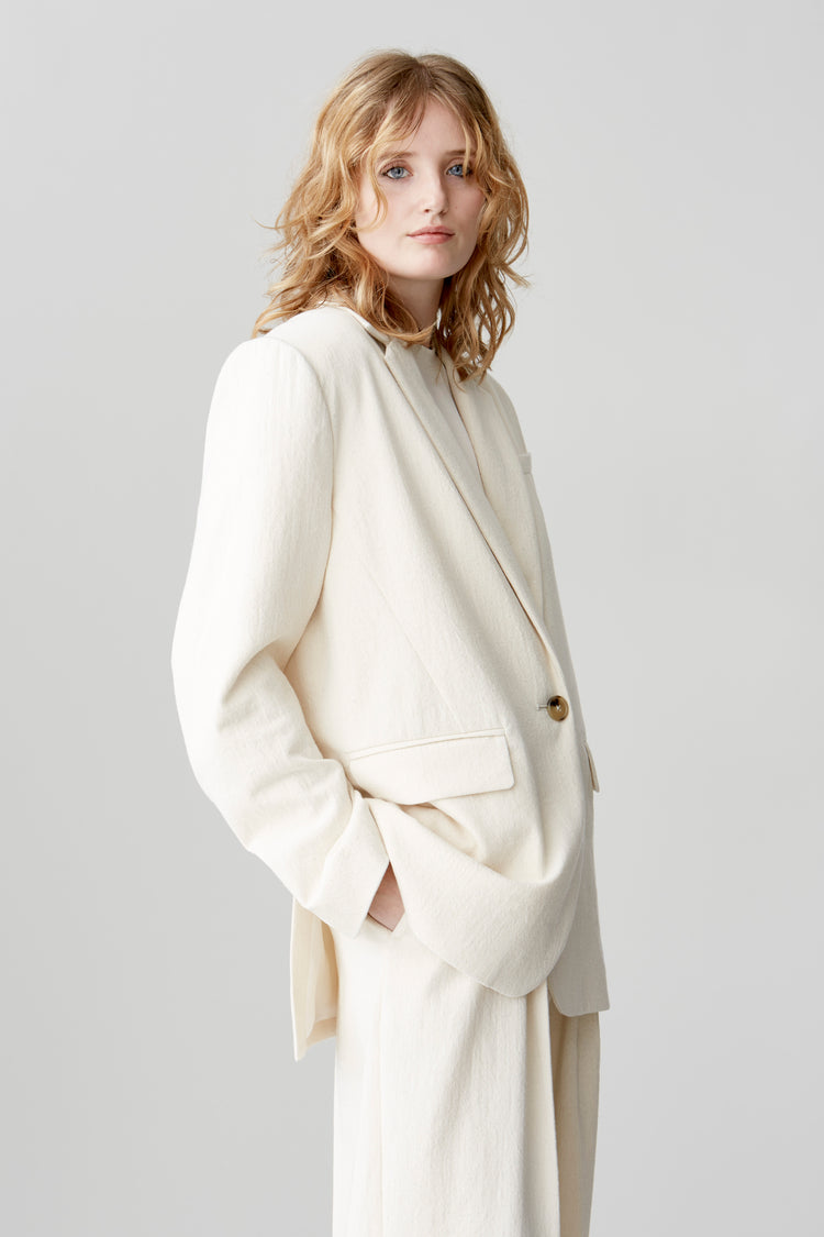 A cropped image of a woman wearing a horn-buttoned, cream-hued, single-breasted jacket styled as a suit with matching pants and a white T-shirt.