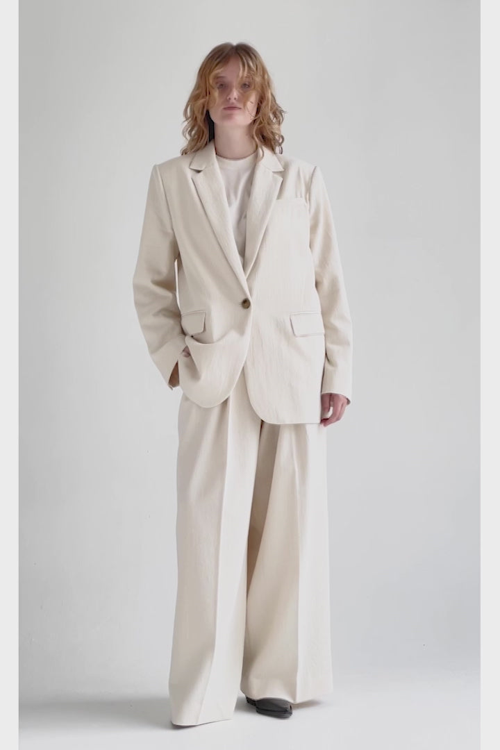 A video of a woman turning around in a cream-hued, wide-legged pair of trousers with pleats putting her hands in side slit pockets, styled as a suit with a matching single-breasted jacket and a white T-shirt. The mid-weight fabrics in the slightly loose-cut suit flow when she moves.